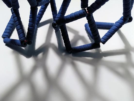 A blue fishing net made of ceramic beads on white background on the upper part of the picture. The bottom part is its shadow. We can't really distinguish when the fishing net stops and the shadow starts. Visible and invisible.