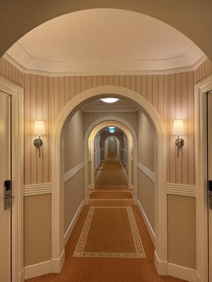 A well-lit hotel corridor featuring multiple archways, plush carpet, and decorative wall sconces. The walls are adorned with vertical stripes, creating a warm and inviting atmosphere.

Ein gut beleuchteter Hotelkorridor mit mehreren Bögen, Plüschteppichen und dekorativen Wandleuchten. Die Wände sind mit vertikalen Streifen geschmückt, was eine warme und einladende Atmosphäre schafft.