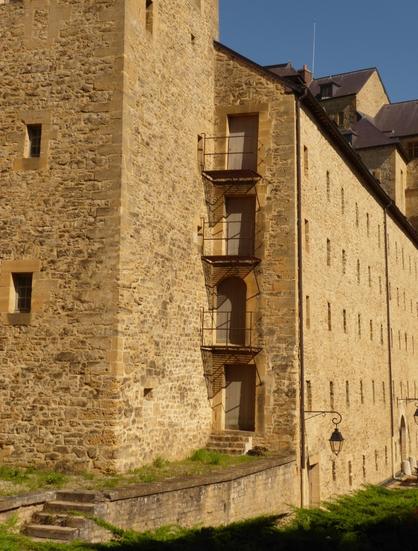 Les fortifications du Château-haut de Sedan en plein soleil sous un ciel dun beau bleu. Avec sa tour et ses petites meurtrières, suivie d'une superposition verticale de portes avec balcon en ferraille et dans son prolongement, une longue façade aux nombreuses petites ouvertures.