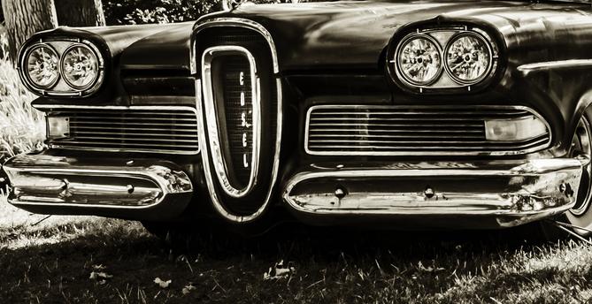 Front view of a vintage Edsel car from the late 1950s, featuring a vertical grille with the name 'EDSEL' spelled out. The car is parked on grass, photographed in black and white with chrome detailing and dual round headlights.