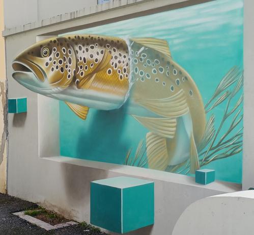 Photographie numérique couleur en format presque carré, avec la vue en perspective d'un mur beige d'où semble surgir la tête et l'avant d'un poisson doré avec des taches noires entourées de blanc et un ventre blanc. Sa queue et ses nageoires, courbées vers la gauche, sont immergées près d'une algue. Des carrés bleus autour de la scène semblent eux aussi être en relief. L'illusion optique est très réussie. Le sol est goudronné.