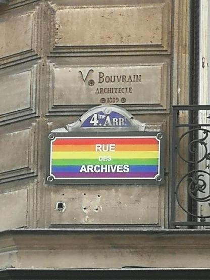 Foto eines Strassenschildes an der Rue des Archives in Paris, das mit den Farben der Regenbogenflagge hinterlegt ist.