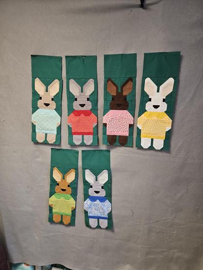 Six sown girl bunnies in different shades of grey and brown, with patterned dresses with solid collars and hems, in light and dark blue, red, pale pink, yellow and green. On a dark green bagground.