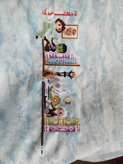 A WIP cross stitch with Mary Shelley and Tolkien sitting on a bookshelf with motif from their books and some books on either side.