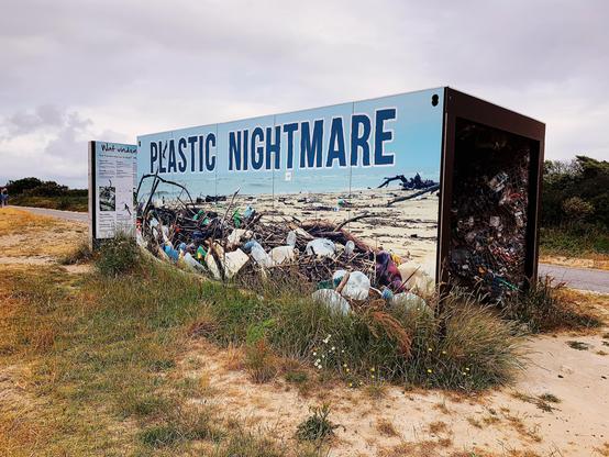 Large outdoor installation featuring a billboard titled "Plastic Nightmare" with a visual of a littered beach. The adjacent compartment is filled with assorted real waste items, mimicking the imagery on the billboard. The installation includes an educational panel on the left, set against a natural landscape with cloudy skies and grassy terrain.