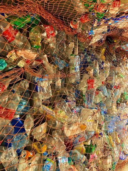 Numerous compressed plastic bottles of various colors, sizes, and brands are tightly packed and secured with an orange mesh net, creating a dense wall of recycled material.