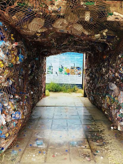 An outdoor tunnel constructed from wire mesh filled with compacted waste like plastic bottles and cans. The interior features netting on the roof section and a large educational poster on marine life at the end of the path. The ground is a wet concrete walkway with scattered small debris.