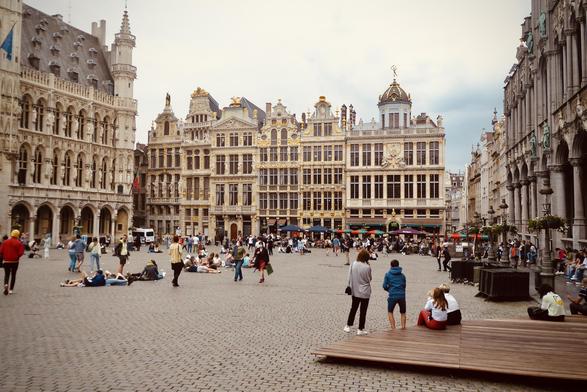 Busy square in a European city, filled with people sitting and walking on cobblestones. The background showcases ornate guild buildings with golden statues and intricate facades, under an overcast sky. In the foreground, several groups engage in casual activities, including sitting on steps and a wooden platform.