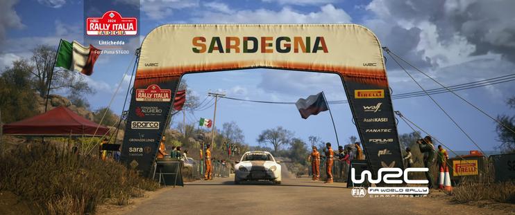 A screenshot from a racing video game, depicting a white Ford Puma S1600 rally car approaching a large start line archway that says "SARDEGNA" in orange letters. The archway is black with sponsor logos. Italian and Sardinian flags are visible. Marshalls in orange suits line the dirt road. A red tent is on the left. In the top left, a rally event logo reads "2024 RALLY ITALIA SARDEGNA."