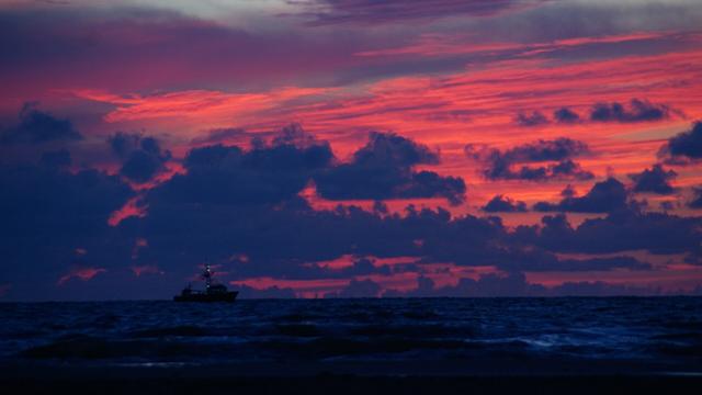 A fishing boat, on the sea, with a background of a read sky, due to sunset, partly covered by blue clouds
