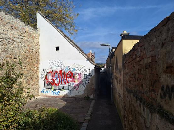 Eine enge Gasse zwischen Häusern und Mauern. Die weiße Hausmauer trägt Graffiti.