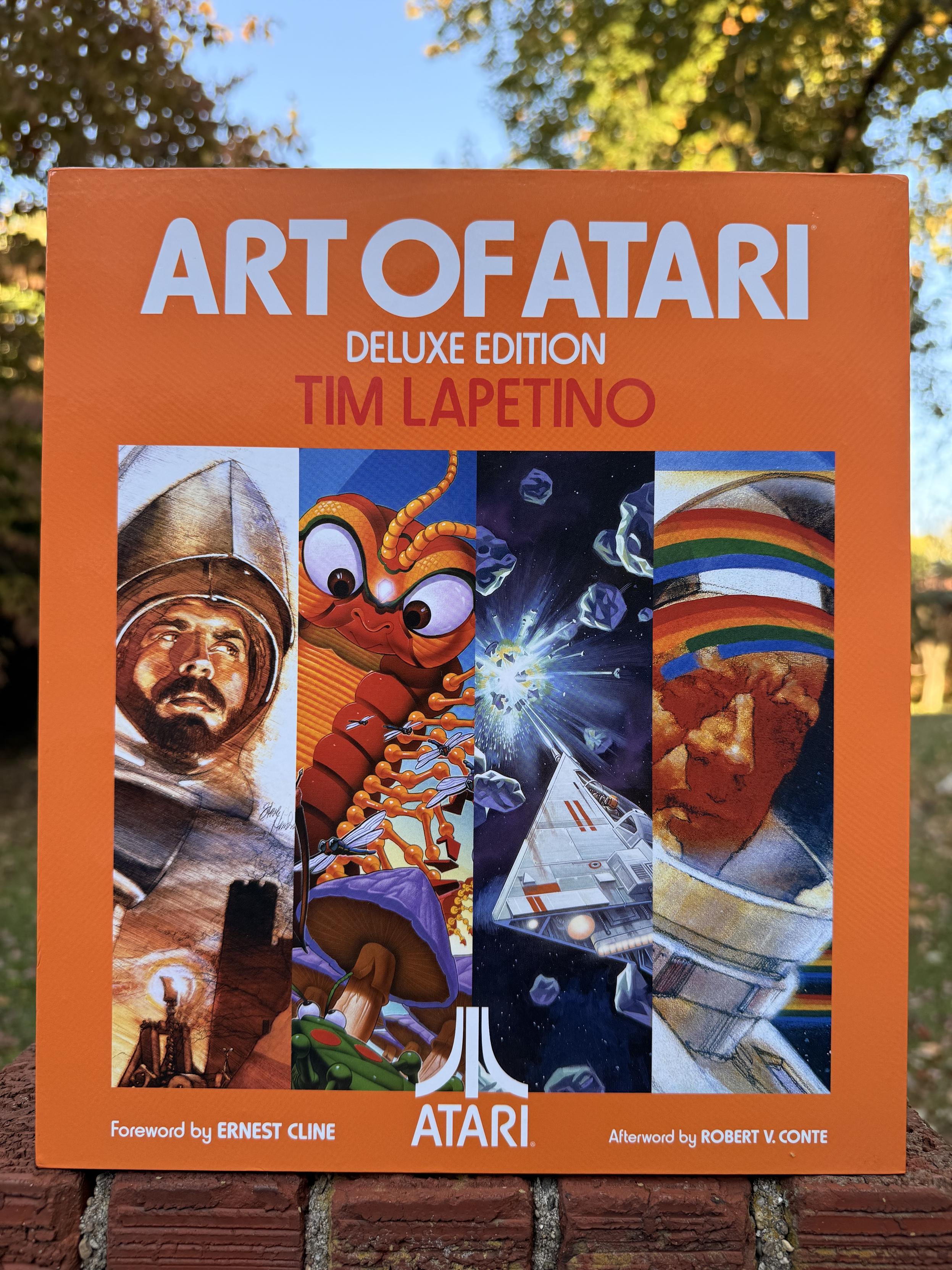 A book titled “Art of Atari”, Deluxe Edition, by Tim Lapetino, Foreward by Ernest Cline, Afterward by Robert V. Conte. The slipcover is orange with white text, and has four vertical panels featuring various examples of Atari game box art.