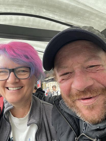 Aufnahme von zwei Personen als Selfie geknipst: rechts im Vordergrund, ein Mann mit Bart und einem dunklen Käppi. Er hat ein freundliches Gesicht und lächelt. Links daneben etwas weiter hinter eine weibliche Person mit Brille und blauen und pinken Haaren, die auch in die Kamera lacht.