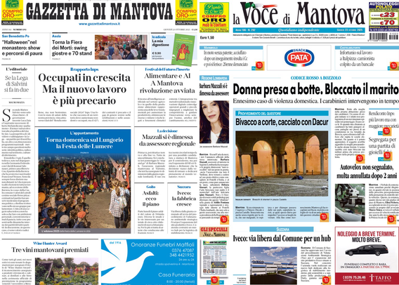 Due prime pagine di quotidiani locali visualizzate su uno schermo in una stanza buia. A sinistra c'è la "Gazzetta di Mantova" e a destra "La Voce di Mantova". I titoli principali riguardano temi di attualità locale, come aumenti dell'occupazione ma con lavori più precari, crisi lavorative, e un caso di violenza domestica con la notizia "Donna presa a botte. Bloccato il marito".