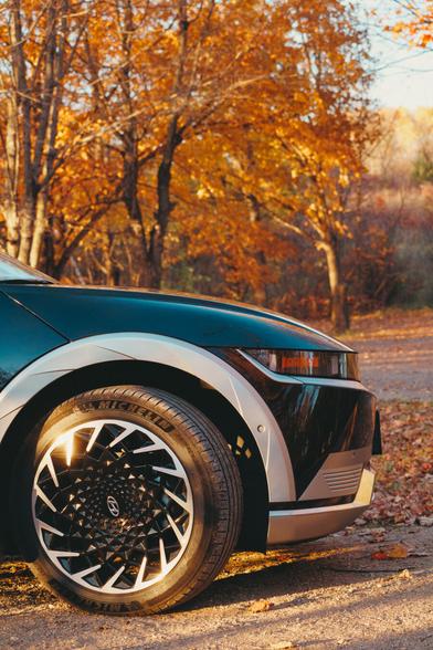 Front view of a Hyundai Ioniq 5 parked on a dirt road surrounded by golden autumn trees. The sunlight catches the car’s geometric wheel design, reflecting warm tones of orange leaves and late afternoon light. The scene feels calm and crisp, capturing both technology and nature in harmony.