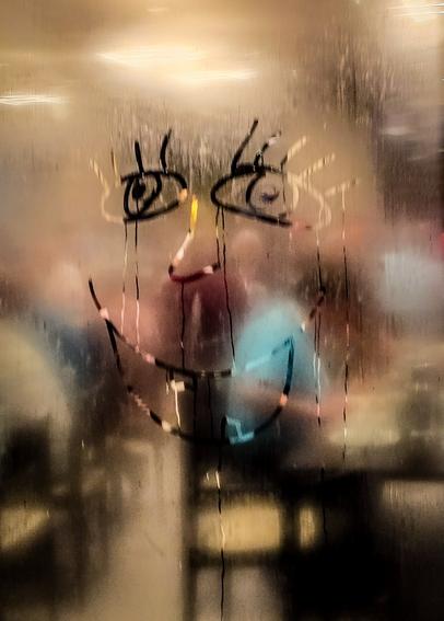 Ein lachendes Gesicht ist mit dem Finger in eine beschlagene Glasscheibe gezeichnet. Durch das Glas schimmern verschwommene bunte Lichter und undeutliche Konturen eines Raums im Hintergrund.