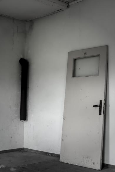 Aufnahme einer schräg an die Wand gelehnten Tür mit der Nummer 15 in der Lagergedenkstätte Sandbostel, daneben ein schwarzes Rohr an der Wand.