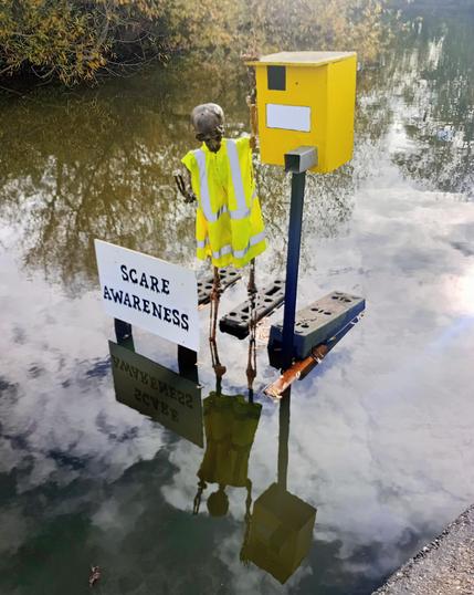 A Halloween skeleton, wearing a high viz vest, with a fake speed camera. It has a sign which reads "scare awareness".