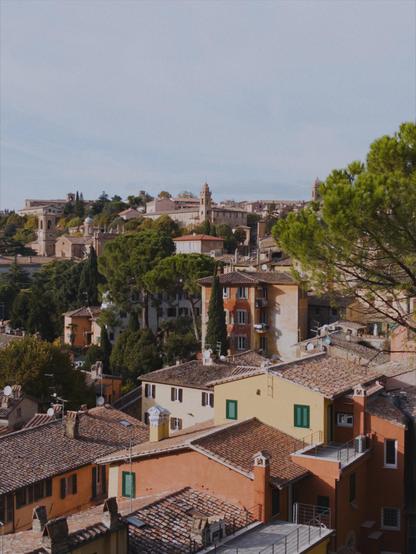A panoramic view of Perugia, showing many old buildings and rooftops over a hill, with pine trees here. The sky is mostly clear with a thin veneer of humidity.
The picture is shot with an iPhone 16e using No Fusion with an AgX LUT.