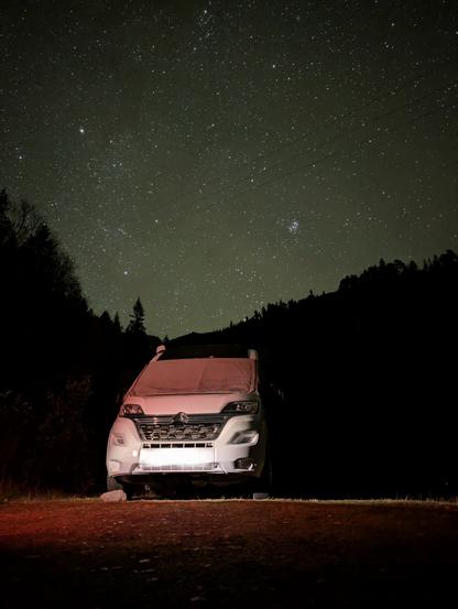 A van with a windshield covered parked in the middle of a forest. The sky is full of stars.