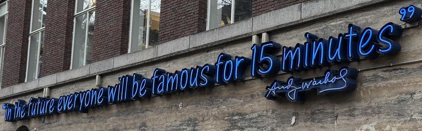 A neon sign displays the text: "In the future everyone will be famous for 15 minutes," attributed to Andy Warhol. The sign is mounted on a brick wall with large windows visible in the background.