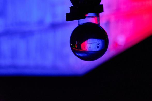 Blick auf eine Glaskugel. In ihr sieht man eine farbig illuminierte Baracke. Das Gebäude ist im Hintergrund unscharf zu erkennen. Das Bild wurde auf den Kopf gestellt.