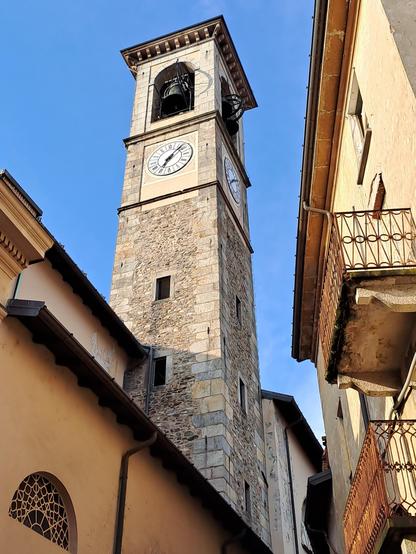 Kirchturm aus hellem Stein mit Uhr und Glocken.
Er ragt in einer schmalen Gasse empor.
Dahinter strahlend blauer Himmel.