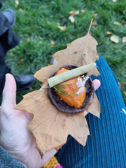 A hand holds a yellow leaf with a small meal of a purple corn cracker, with creamed pumpkin, dill and half a green bean.