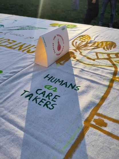 A painted linen table cloth that says in green letters ' Humans as Care Takers'. A small upright sign casts a shadow, with pumpkin printed on it, in a circle saying ' The Vegetable Parliament Part#5'
