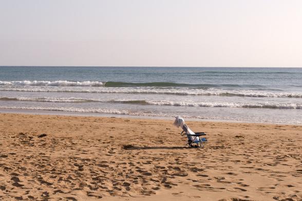 A serene beach scene with a single lounge chair placed slightly off-center on warm, beige sand, its surface smooth and subtly textured by wind and light. A soft white towel drapes over the chair, catching the sunlight and adding a gentle contrast to the neutral tones. In the background, pale blue waves roll calmly toward the shore beneath a clear, pastel sky, creating a rhythmic horizon line. The composition is minimal and balanced, while the interplay of soft colors and textures evokes a tranquil, contemplative atmosphere—an invitation to solitude by the sea.