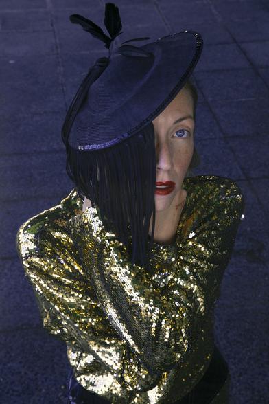 Sicut wearing a shiny gold sequined dress and a black hat with fringe and feathers, partially covering their face. They have bright red lipstick and are standing on a tiled sidewalk, looking directly at the camera with a dramatic, artistic expression on their face.