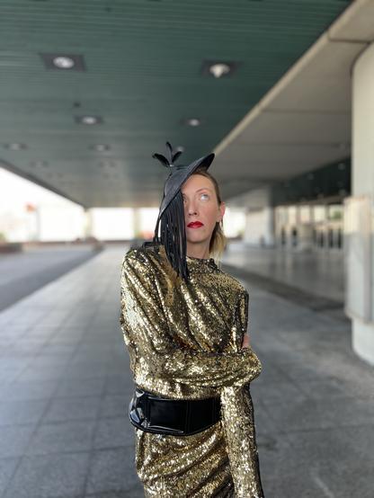 Sicut wearing a gold sequin dress with a wide black belt and a black fascinator hat featuring fringe and feather details. They have red lipstick and are posing with one arm crossed under the other in an urban setting with concrete columns and a green ceiling overhead. Sicut is rolling her eyes up.