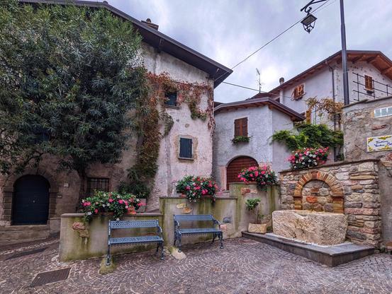 A charming, rustic courtyard in what appears to be a quaint European village. The scene features a cobblestone pavement that adds to the old-world charm of the setting. Two vintage-style metal benches are positioned against a low stone wall, which is adorned with vibrant flower pots filled with blooming pink flowers.

The buildings surrounding the courtyard are constructed from a mix of stone and stucco, with wooden shutters on the windows and climbing plants adding a touch of greenery. One of the buildings is partially covered in creeping vines, enhancing the picturesque quality of the scene.

In the centre-right of the courtyard, there is a small stone structure, possibly an old well or fountain, contributing to the historical ambiance. The sky above is overcast, casting a soft, diffused light over the tranquil setting. The overall atmosphere is peaceful and inviting, evoking a sense of timelessness and simplicity.