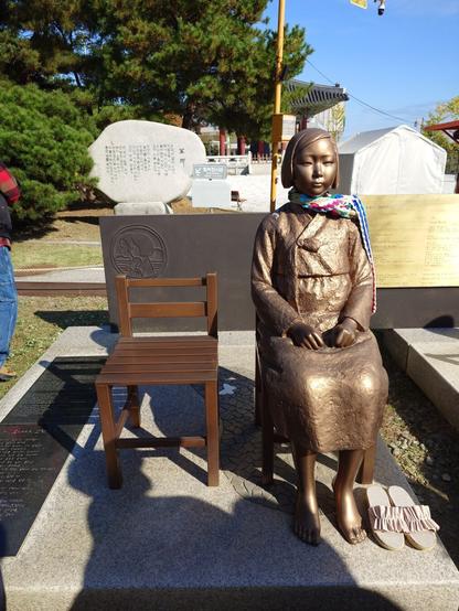 Monument to the comfort ladies. A young girl made of bronze sitting on a chair. the chair next to her is empty.
