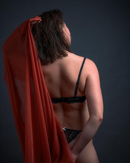 Woman with shoulder-length dark hair facing away, wearing black lace lingerie. She holds a sheer red fabric draped over her left shoulder. Dark backdrop enhances contrast with her skin and clothing.