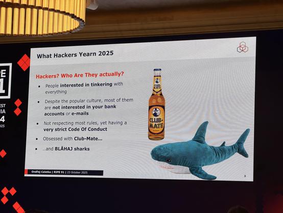 A photo of a slide diplayed on the RIPE 91 conference.
The slide has a picture of a Club Mate bottle and a BLAHAJ shark from Ikea. The title of the slide "What Hackers Yearn 2025" refers to the WHY2025 conference. The text reads:
Hackers? Who Are They actually?
- People interested in tinkering with everything
- Despite popular culture, most of them are not interested in your bank accounts or e-mails
- Not respecting most rules, yet having a very strict Code of Conduct
- Obsessed with Club-Mate
- and BLÅHAJ sharks

Slide is signed by Ondřej Caletka