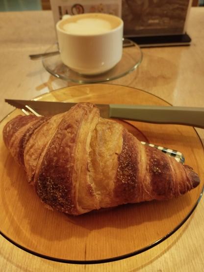 Imagen de un desayuno en una mesa de madera clara. En primer plano, un croissant grande y dorado, de apariencia crujiente y hojaldrada, reposa sobre un plato redondo de cristal ámbar. A su lado, se observa un cuchillo con mango verde claro. Detrás del croissant, se ve una taza blanca de café con leche, adornada con arte latte en forma de corazón o hoja en su espuma cremosa. La taza está sobre un platillo de cristal transparente.
