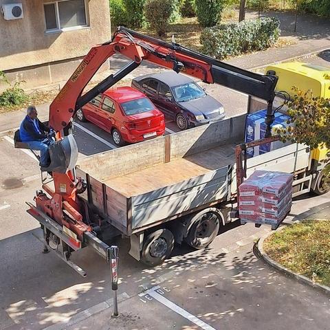 This image shows an overhead view of a truck with a crane attachment (a knuckle-boom loader, specifically a Palfinger model) being operated by a man. The crane is lifting a pallet of red and white bags (labeled Baumit, likely construction materials) from the back of the truck. Two parked cars, one red and one dark gray, are visible behind the crane in an asphalt parking area. A yellow truck or container is partially visible on the right.