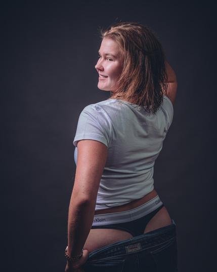 Woman with wet hair is standing in a casual pose against a neutral dark background, looking over her shoulder. She is wearing a white t-shirt and Calvin Klein underwear, with low-rise jeans being partially pulled down. Her left hand is resting on her hip, and she has a slight smile on her face.