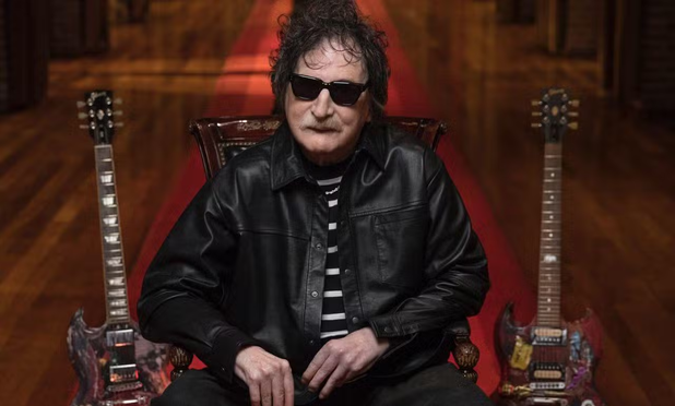 Charly Garcia está sentado en el centro de la imagen, llevando una chaqueta de cuero negra sobre una camiseta a rayas horizontales blancas y negras. Tiene el pelo oscuro y rizado, un bigote y usa gafas de sol oscuras que cubren sus ojos. A cada lado del hombre, hay una guitarra eléctrica de cuerpo oscuro y diseño personalizado, apoyada verticalmente. El fondo es oscuro con iluminación cálida que proyecta un brillo rojizo y anaranjado, sugiriendo un escenario o estudio.
