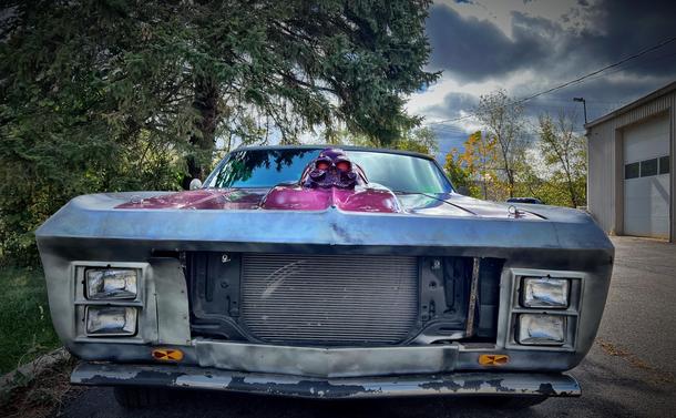 A front view of a customized car featuring a distinctive purple paint job and a skull ornament on the hood. The car has an unfinished look, with exposed parts and a cloudy sky in the background. It is likely a mid-1960s Chevy Impala