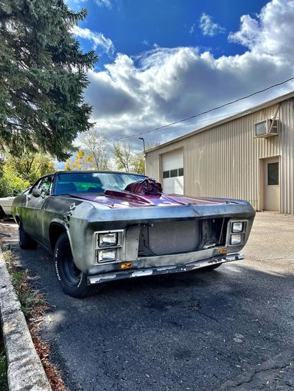 A partially restored vintage car with a metallic silver body and a maroon hood is parked near a building. The setting features a cloudy blue sky and a nearby tree, with hints of fall foliage in the background. It is likely a mid-1960s Chevy Impala