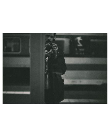 black and white photograph of myself in front of a mirror. very dark and grainy photo. on the left side you see a train on the background platform, in the middle I'm looking with my camera and headphones around a vertical metal construction that is cutting me in half, followed by the mirror reflection an other train on the right hand side of the photo.