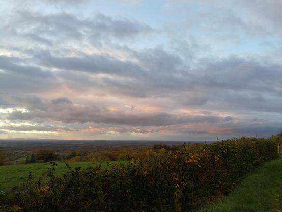 Vue sur la plaine de Bresse à la tombée de la nuit. Une haie au premier plan, puis une prairie, au loin des bosquets d'arbres feuillus. Le ciel est encore bien nuageux suite au passage des volutes de la tempête Benjamin, et les nuages vont du gris au rose