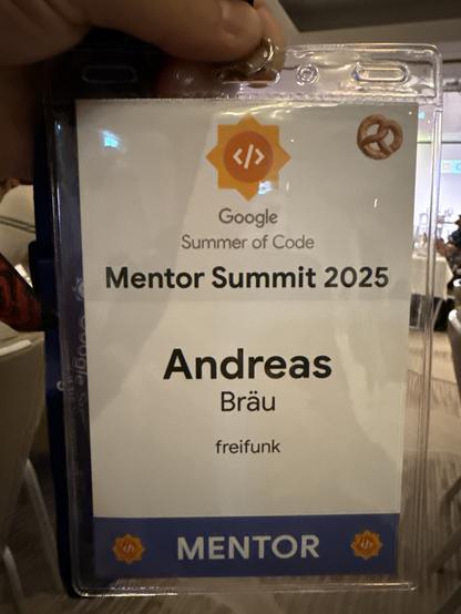 A name badge for the Google Summer of Code Mentor Summit 2025, held by a person. It features the name "Andreas Bräu," the designation "MENTOR," and the organization "freifunk." The badge includes a pretzel.