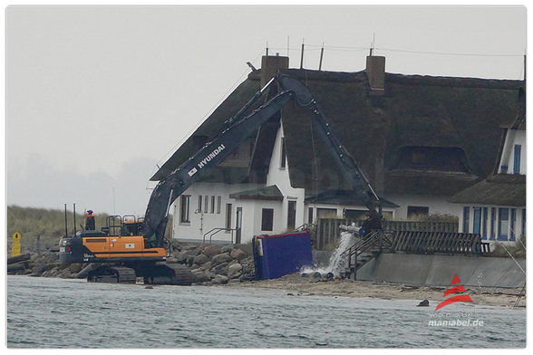 Bagger steht mit den Ketten im Ostseewasser vor einem Haus und lädt Buhnen ab. Ostseewasserdusche gibt es gratis.