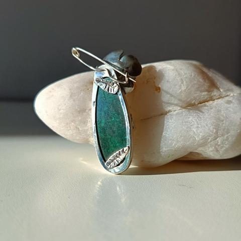the back side of a silver lapel pin with a big green gemstone. We can see two small silver leaves an the pin on top. it's placed next to white pebble