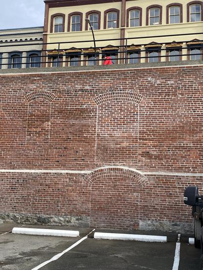brick wall of a ruined warehouse with bricked in windows and wagon entry, above an asphalt parking lot and below 1860s commercial frontage on the other side of the street