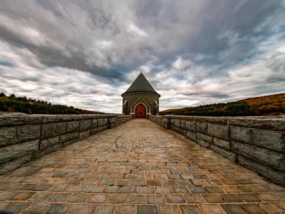 A stone walkway with short stone walls on both sides. A the end of walkway is cylindrical building with conical shaped roof and large arch wood door at the center. The clouds about of the sky are grey and very dramatic