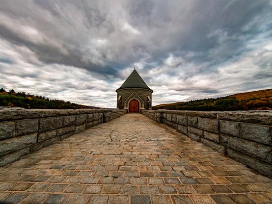 A stone walkway with short stone walls on both sides. A the end of walkway is cylindrical building with conical shaped roof and large arch wood door at the center. The clouds about of the sky are grey and very dramatic